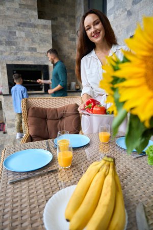 Téléchargez les photos : Belle femme met la table sur la terrasse, papa et fils adolescent préparent la nourriture dans la zone barbecue - en image libre de droit