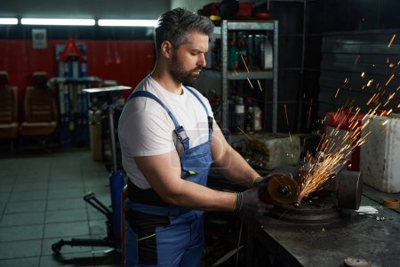 Seriöser Kfz-Mechaniker in Arbeitskleidung schleift metallene Autoteile in Schraubstock in Werkstatt eingeklemmt