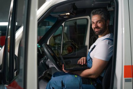 Téléchargez les photos : Smiling technicien de service automobile assis dans la voiture du client avec ordinateur portable dans les mains - en image libre de droit