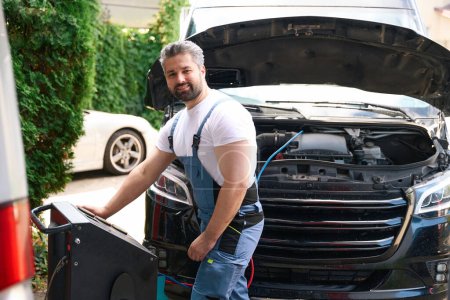 Téléchargez les photos : Joyeux technicien de service automobile rechargeant le système de climatisation de voiture client avec l'équipement - en image libre de droit