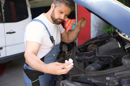 Téléchargez les photos : Serieux concentré technicien auto essuyant jauge avec chiffon devant le capot de voiture ouverte - en image libre de droit