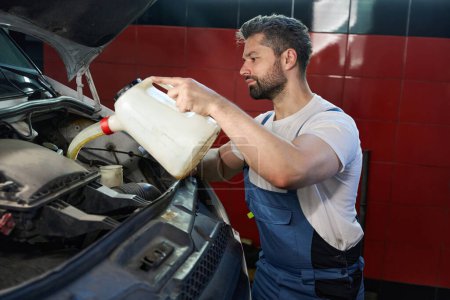 Photo for Concentrated automotive technician pouring motor oil from plastic tank into car engine while standing in front of open vehicle hood - Royalty Free Image