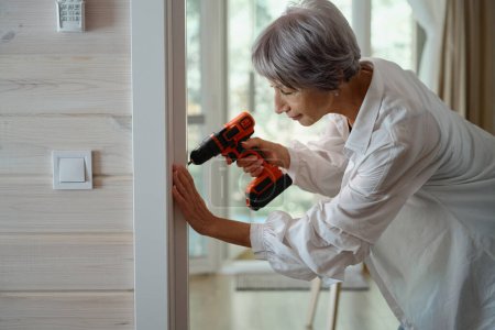 Téléchargez les photos : Femme âgée fait des réparations mineures maison sur son propre, elle utilise un tournevis électrique - en image libre de droit