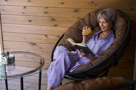 Foto de Mujer mayor se sienta en una silla acogedora en la terraza con un libro y un vaso de jugo - Imagen libre de derechos