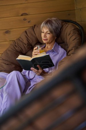 Foto de Elegante anciana se sienta en una silla acogedora en la terraza con un libro y un vaso de jugo - Imagen libre de derechos
