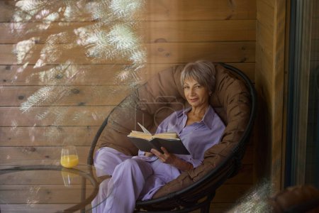 Foto de Mujer elegante se sienta en una silla acogedora en la terraza con un libro y un vaso de jugo - Imagen libre de derechos