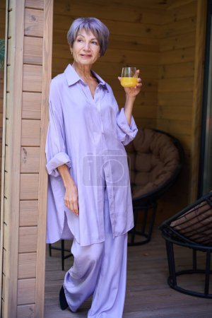 Téléchargez les photos : Spectaculaire dame âgée se dresse sur une terrasse en bois avec un verre de jus, il y a des meubles de campagne sur la terrasse - en image libre de droit