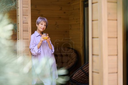 Téléchargez les photos : Spectaculaire dame se dresse sur une terrasse en bois avec un verre de jus, il ya des meubles de campagne sur la terrasse - en image libre de droit