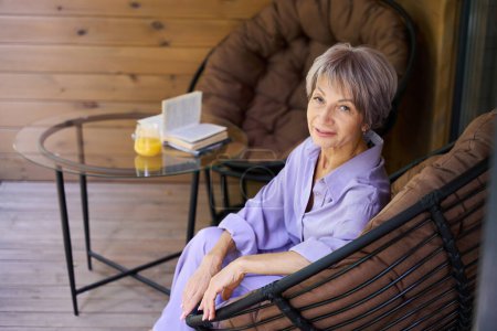 Téléchargez les photos : Belle femme assise dans une chaise confortable sur une terrasse en bois, à côté d'un livre et un verre de jus - en image libre de droit