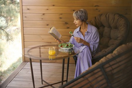 Foto de Mujer mayor con ropa lila se sienta en la terraza con un libro y comida ligera - Imagen libre de derechos