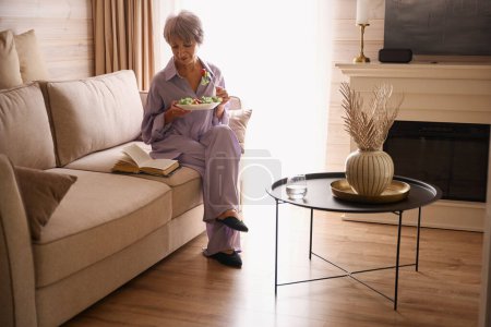Téléchargez les photos : Vieille dame mignonne installée sur un canapé confortable avec un livre et de la nourriture légère, femme en vêtements lilas - en image libre de droit