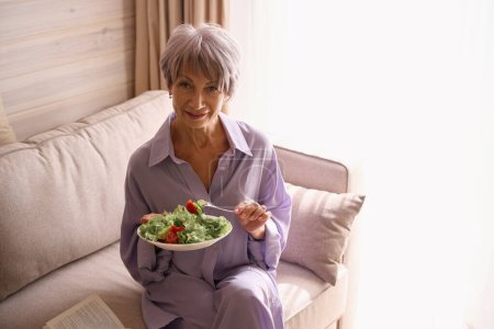 Foto de Linda anciana sentada cómodamente en un sofá acogedor con un libro y comida ligera, mujer en ropa lila - Imagen libre de derechos