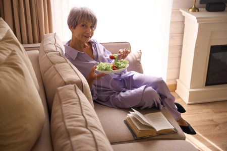 Foto de Encantadora dama se sienta cómodamente en un acogedor sofá con un libro y comida ligera, una mujer en ropa lila - Imagen libre de derechos
