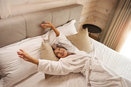 Téléchargez les photos : Heureuse femme âgée se réveille dans une chambre confortable sur des oreillers doux, elle s'étend doucement sur le lit - en image libre de droit