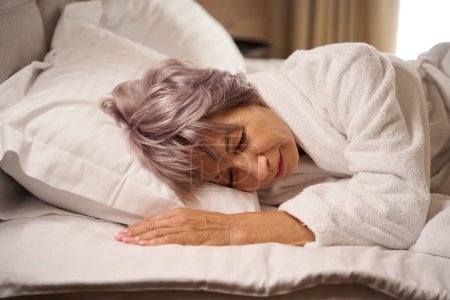 Téléchargez les photos : Femme âgée dort doucement dans une chambre lumineuse sur des oreillers doux, elle est dans un peignoir doux - en image libre de droit
