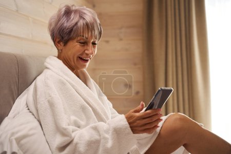 Foto de Señora feliz en un albornoz se sienta en la cama con un teléfono móvil, ella se sienta en almohadas suaves - Imagen libre de derechos