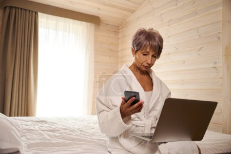 Téléchargez les photos : Pensionnée communique sur son téléphone portable et utilise un ordinateur portable, elle s'assoit sur le lit dans la chambre - en image libre de droit