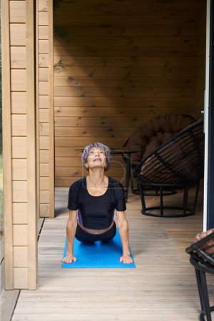 Téléchargez les photos : Femme âgée fait du yoga sur une terrasse en bois confortable, elle utilise un tapis de rouleau - en image libre de droit