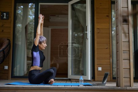 Téléchargez les photos : Femme en tenue de sport fait du yoga devant un ordinateur portable, elle est située sur une terrasse en bois - en image libre de droit