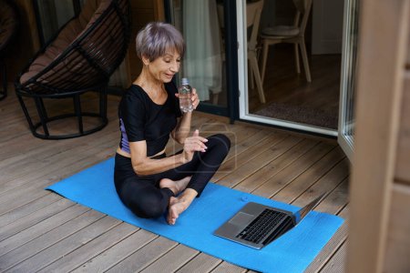 Photo for Pleasant woman in sportswear communicates online, she sits on a roll mat on the terrace - Royalty Free Image