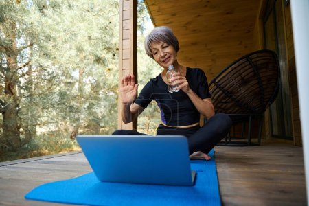 Téléchargez les photos : Femme âgée en vêtements de sport communique en ligne, elle est assise sur un tapis roulant sur la terrasse - en image libre de droit