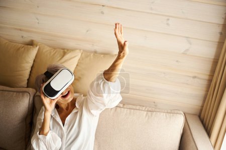 Foto de Feliz pensionista se sienta en el sofá en la habitación jugando un juego virtual, ella utiliza gafas de realidad virtual - Imagen libre de derechos