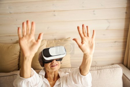 Téléchargez les photos : Femme âgée passe son temps libre sur le canapé à jouer à un jeu virtuel, elle utilise des lunettes de réalité virtuelle - en image libre de droit
