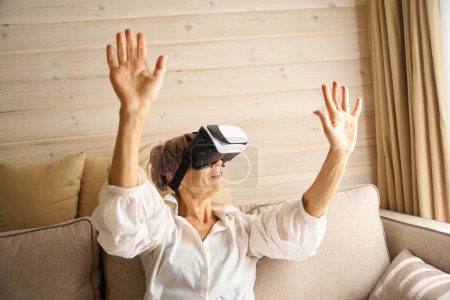 Foto de Mujer mayor pasa su tiempo libre en el sofá jugando un juego virtual, ella utiliza gafas de realidad virtual - Imagen libre de derechos