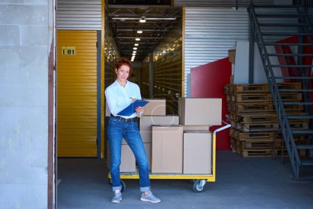 Photo for Smiling woman standing with a blue folder near a cargo trolley, she is in casual clothes - Royalty Free Image