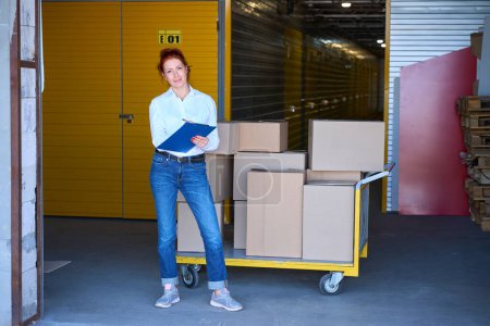 Photo for Red-haired woman stands with a blue folder near a cargo cart, she is in casual clothes - Royalty Free Image