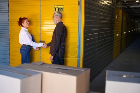 Photo for Female with a blue folder shakes hands with a bearded man, people are in a warehouse - Royalty Free Image
