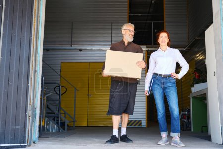 Téléchargez les photos : Une femme avec un dossier bleu et un homme sont dans un entrepôt, l'homme a une boîte en carton - en image libre de droit