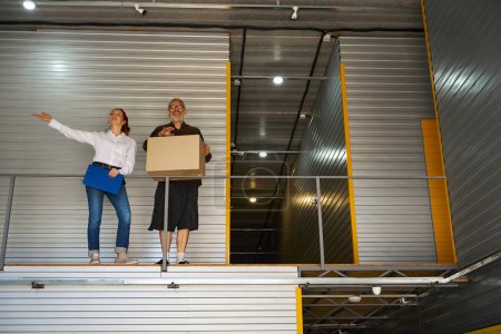 Foto de Mujer con carpeta azul y hombre están en el segundo piso del almacén, el hombre tiene una caja de cartón - Imagen libre de derechos