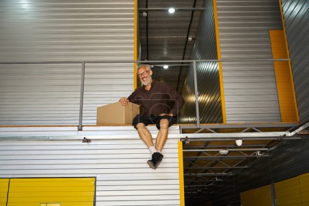 Foto de Alegre hombre sentado en un contenedor con una caja de cartón, colgó sus piernas - Imagen libre de derechos