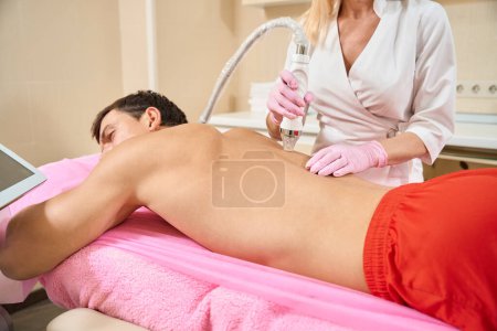 Foto de Doctor en cosmetología clínica hace RF levantamiento de la piel de la espalda de un joven, un cliente en pantalones cortos de color naranja - Imagen libre de derechos