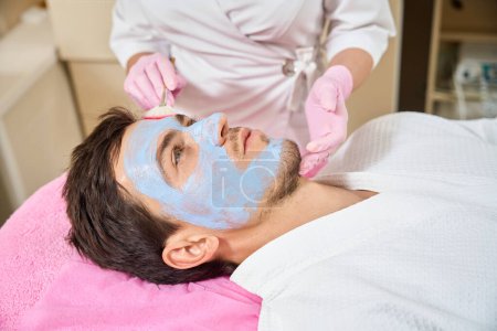 Photo for Brown-haired man lies on a comfortable couch in a cosmetology office, a cosmetologist applies a cleansing mask to his face - Royalty Free Image
