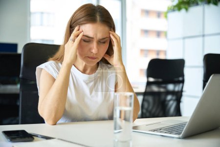 Photo for Female suffers from a headache at the workplace, in front of her is a laptop and a glass of water - Royalty Free Image