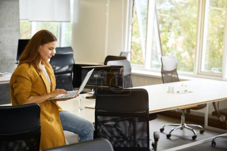 Téléchargez les photos : Femme travaille avec un ordinateur portable dans un bureau de coworking, elle s'assit sur le bord de la table de bureau - en image libre de droit