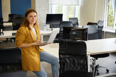 Photo for Manager is working with laptop in the coworking office area, she sat down on the edge of the office table - Royalty Free Image