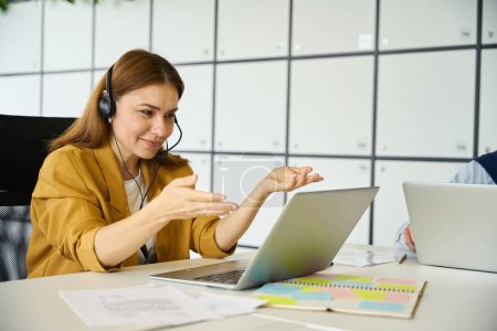 Téléchargez les photos : Jeune gestionnaire communique en ligne dans un bureau de coworking, elle utilise un ordinateur portable et un casque - en image libre de droit
