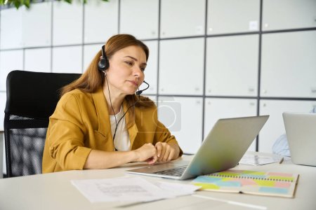 Téléchargez les photos : Femme gestionnaire fatiguée communique en ligne dans un bureau de coworking, elle utilise un ordinateur portable et un casque - en image libre de droit