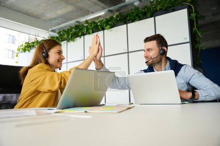 Foto de Hombre y una mujer se saludan en una gran mesa de oficina, la gente está usando computadoras portátiles y auriculares - Imagen libre de derechos
