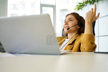 Photo for Young manager is working at a desk in a coworking office area, she is using a laptop and a headset - Royalty Free Image