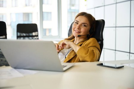 Photo for Young woman working at a desk in a coworking office area, she is using a laptop and headset - Royalty Free Image
