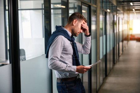 Téléchargez les photos : L'homme se tient dans un couloir de bureau avec des documents de travail, il souffre d'un mal de tête - en image libre de droit