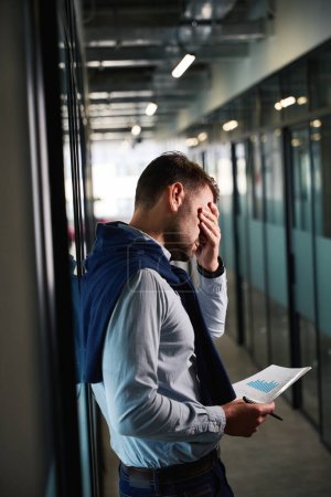 Téléchargez les photos : L'homme fatigué se tient dans un couloir de bureau avec des documents de travail, il souffre d'un mal de tête - en image libre de droit