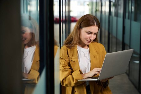 Téléchargez les photos : Belle femme debout avec un ordinateur portable dans le couloir d'un espace de coworking, elle travaille dans un environnement démocratique - en image libre de droit