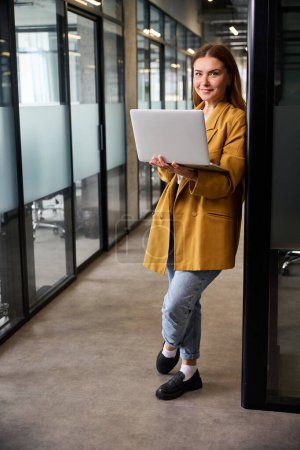 Téléchargez les photos : Femme pigiste debout avec un ordinateur portable dans le couloir d'un espace de coworking, elle travaille dans un environnement démocratique - en image libre de droit