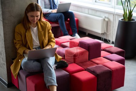 Téléchargez les photos : Femme assise avec un ordinateur portable sur un canapé multicolore, sa collègue travaille à proximité - en image libre de droit