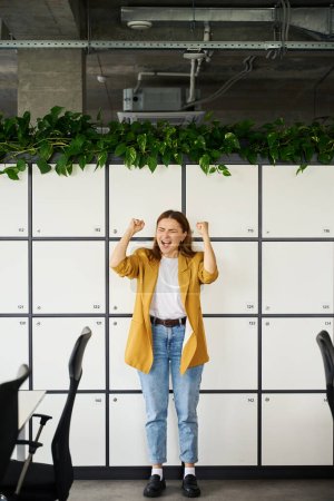 Téléchargez les photos : Femme pigiste en veste jaune se réjouit dans la zone de bureau d'un espace de coworking, elle se tient près des casiers de stockage - en image libre de droit